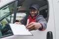 Positive African-American deliveryman behind the wheel of white delivery truck showing documents to sign and a pen to Royalty Free Stock Photo