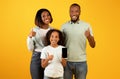 Positive african american family of three demonstrating smartphone and showing thumbs up over yellow background Royalty Free Stock Photo