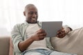 Positive african american mature man spending time with digital tablet, browsing internet while resting on comfy couch Royalty Free Stock Photo