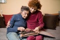 Positive aged ladies looking album photos sitting sofa at home, cheerful friends. Senior woman and her mature nurse watching photo