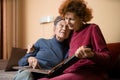 Positive aged ladies looking album photos sitting sofa at home, cheerful friends. Senior woman and her mature nurse watching photo