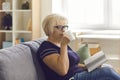 Positive aged woman grandma in t-shirt and glasses sitting on sofa with book and drinking tea