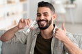 Positive Arab guy drinking fresh clear water from glass, showing thumb up gesture and smiling at camera at home