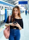 Positive Asian female standing on platform at railroad station and browsing mobile phone while looking at camera. Idea for