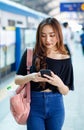 Positive Asian female standing on platform at railroad station and browsing mobile phone while looking at screen. Idea for