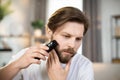 Positive attractive young guy with beard trimming his beard on his cheek with an electronic razor. Royalty Free Stock Photo