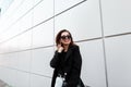 Positive attractive young hipster woman in stylish dark sunglasses in an elegant black coat with a leather bag in jeans posing Royalty Free Stock Photo