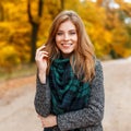 Positive attractive young woman in a warm gray fashionable modern outerwear with a green scarf in the park. Cheerful girl model