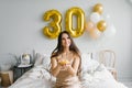 Positive beautiful Caucasian woman is sitting on the bed in the morning and holding a cake with a candle in her hands and smiling Royalty Free Stock Photo