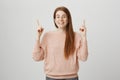 Positive beautiful redhead european woman pointing up with index fingers while smiling broadly and glancing at camera Royalty Free Stock Photo