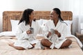 Positive black girlfriends in bathrobes sitting on bed, talking Royalty Free Stock Photo