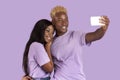 Positive black guy taking selfie with his beloved girlfriend on smartphone over lilac studio background