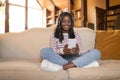 Positive black lady in headphones sitting cross legged on couch, listening to music or audio book on cellphone at home