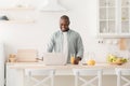 Positive black mature man reading online news on laptop and drinking orange juice in kitchen, enjoying morning at home Royalty Free Stock Photo