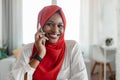 Positive black woman in hijab talking on mobile phone, having pleasant conversation with client, sitting at home office Royalty Free Stock Photo