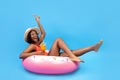 Positive black woman sitting on inflatable ring, holding tropical cocktail on blue background, full length portrait