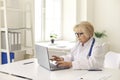 Mature woman doctor in white uniform sitting and working online on laptop Royalty Free Stock Photo