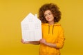 Positive bright woman with fluffy curly hairstyle in hoody looking at paper house with thoughtful expression