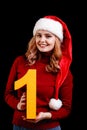 Smiling pretty girl in a christmas hat holding number 1 on a black background. New year 2018 concept. Royalty Free Stock Photo