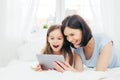 Positive cheerful small female kid and her young mother watch interesting cartoon on tablet, connected to wireless internet, pose