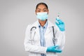 Positive confident young black lady doctor in white coat, protective mask show syringe, recommends vaccination