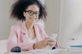 Positive curly haired woman wears spectacles, formal wear, holds modern cell phone, searches information about business news, sits