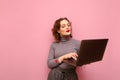 Positive curly redhead girl isolated on pink background with laptop in hands, looks at computer screen. Cute lady browsing the Royalty Free Stock Photo