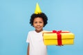 Positive cute happy little boy celebrating birthday, wearing party cone on his head and holding big gift box Royalty Free Stock Photo