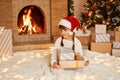 Positive cute little girl wearing white sweater and santa claus hat, holding stack of presents in hands, sitting on floor on soft Royalty Free Stock Photo