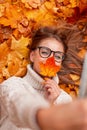 Positive cute young hipster woman holding an orange leaf near face and makes selfie on smartphone. Happy girl in a knitted sweater Royalty Free Stock Photo