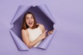 Positive delighted excited brunette woman breaking through purple paper hole wearing casual white top pointing away at copy space Royalty Free Stock Photo