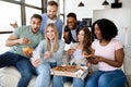 Positive diverse friends having home party, sitting on couch with pizza, chips and smartphone, having fun together