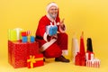 Positive elderly man with gray beard wearing santa claus costume sitting near wrapped gift boxes, pointing to camera and giving Royalty Free Stock Photo