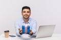 Positive elegant businessman sitting in office workplace, giving gift box to camera and smiling friendly