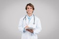 Positive european man doctor therapist in white coat chatting on tablet, smiling at camera isolated on grey background