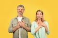 Positive middle aged couple with closed eyes making gesture of gratitude, putting hands to chest, yellow background