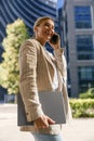 Positive female freelancer holding laptop and talking phone standing on modern building background Royalty Free Stock Photo