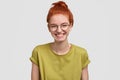 Positive freckled pupil has red hair, smiles broadly, wears round optical glasses and green t shirt, stands against