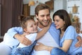 Positive friendly young parents with smiling little daughter sitting on sofa together while relaxing at home on weekend