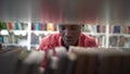 Positive fussy African American man choosing book in library standing near rack with textbooks