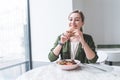 Positive girl eats a sandwich and a salad in a cafe with a light interior and smiles. A look at the camera