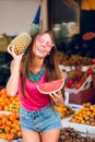 Positive girl with a great smile holding ananas and slice of watermelon on market on fruits background