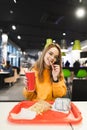 Positive girl holding a glass with a drink in hand, eating french fries, looks at the camera and smiling