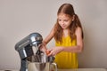 A positive girl of 9-10 years cooks homemade cake in the kitchen, beats eggs in a mixer on the kitchen table. Against