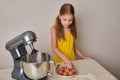 A positive girl of 9-10 years cooks homemade cake in the kitchen, beats eggs in a mixer on the kitchen table. Against