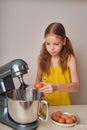 A positive girl of 9-10 years cooks homemade cake in the kitchen, beats eggs in a mixer on the kitchen table. Against