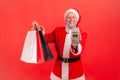 Positive gray bearded man in eyeglasses and santa claus costume holding and showing paper shopping bags and payment terminal,