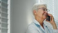 Positive grey haired mature lady doctor with glasses talks on mobile phone with family member near window