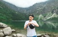 Positive guy in a cap and white t-shirt stands on the shores of Lake Morskie Oko and opens a can with a drink on the background of