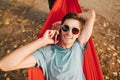 Positive guy in sunglasses lies in a hammock on a tree in the woods, talking on the phone and looking at the camera with a smile Royalty Free Stock Photo